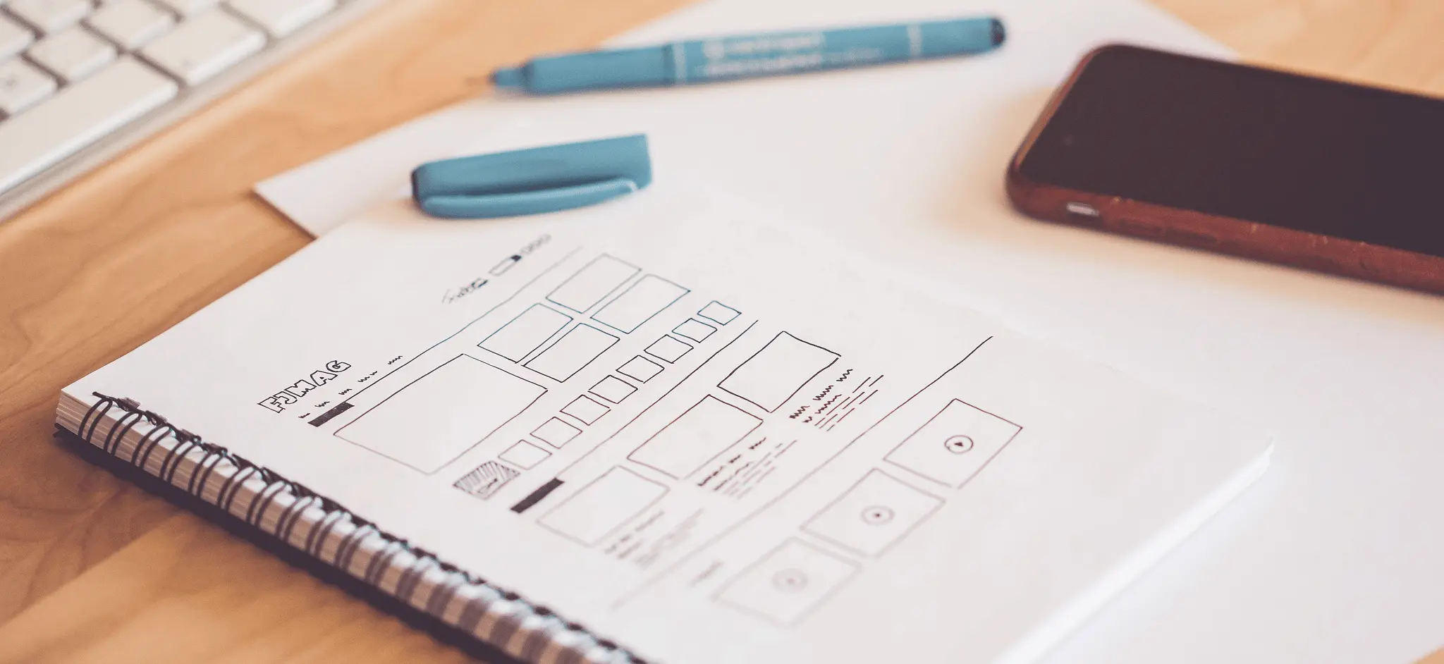 A spiral notebook of hand-drawn website wireframe sketches next to a laptop keyboard and phone on a wooden desk.