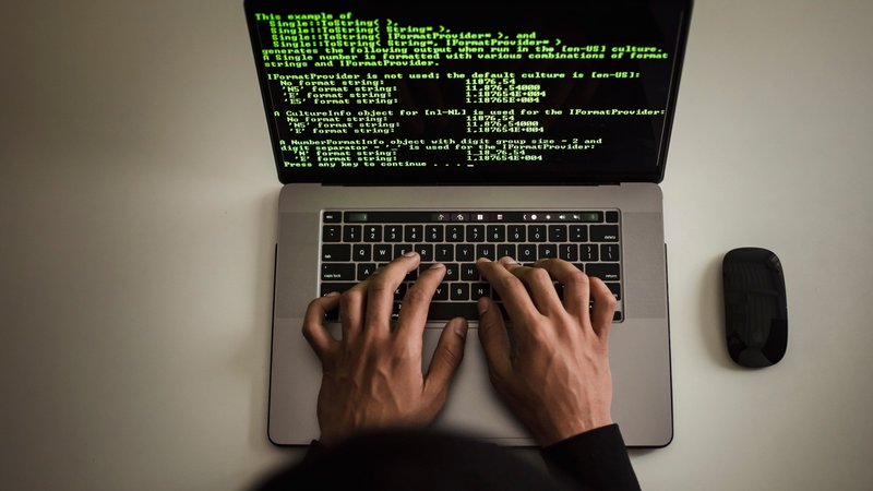 Overhead view of hands typing on a laptop, its screen filled with green code on a black background.